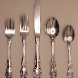 May include: A set of five silver-colored flatware pieces arranged on a neutral background. The set includes two forks, a knife, and two spoons. Each piece has a decorative floral pattern on the handle.