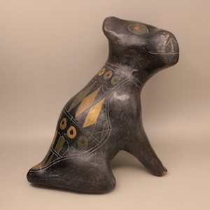 May include: A dark gray ceramic sculpture of a seated animal, possibly a dog or cat. The sculpture has decorative geometric patterns in gold, green, and white. The animal's eyes are accented with green. The sculpture is on a neutral background.