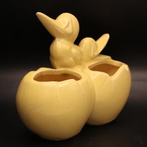 May include: A yellow ceramic duckling planter. The planter is shaped like two cracked eggs with a duckling perched on top. The duckling has a long beak and is facing upwards. The planter is a light yellow color.