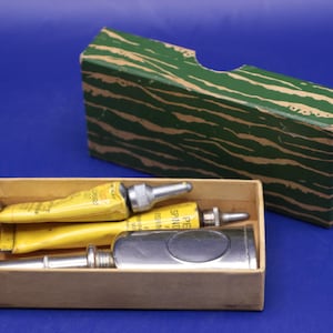 May include: A set of vintage tools, including yellow tubes and a silver tool, presented in a cardboard box. A green and brown patterned box sits behind the open box. The background is a solid blue.