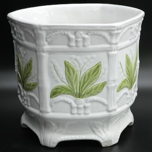 May include: White ceramic planter with a geometric design. The planter features green leaf accents and bamboo-style detailing. The planter has a square shape with a decorative base.