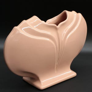 May include: A peach-colored ceramic vase with a unique, sculptural design. The vase has a flared base, a rounded body, and a top with a petal-like opening. The surface is smooth and glossy, reflecting light. The overall shape is organic and modern.