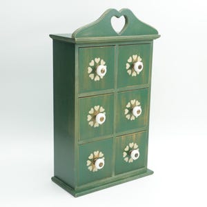 May include: A green wooden cabinet with six drawers, each adorned with a white floral design and a white knob. The top of the cabinet features a heart-shaped cutout. The cabinet is painted in a distressed style.