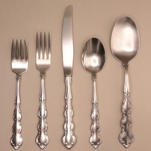 May include: A set of five silver-colored flatware pieces arranged on a neutral background. The set includes two forks, a knife, and two spoons. Each piece has a decorative, ornate handle.