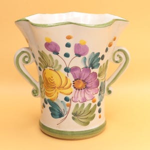 May include: A white ceramic vase with a scalloped rim and two handles. The vase is adorned with hand-painted floral designs in yellow, purple, and green. The base and rim are trimmed with green paint, creating a decorative accent.