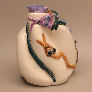 May include: A cream-colored ceramic pitcher featuring a glossy finish and three-dimensional sculpted creatures. The vessel is adorned with a purple, orange, and dark green creature wrapped around the exterior, each with distinct dark eyes.