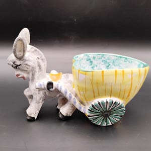 May include: A ceramic donkey pulling a cart planter. The donkey is gray and white with black hooves and a red mouth. The cart is yellow with green and white accents and a turquoise interior. The wheel is green and black.