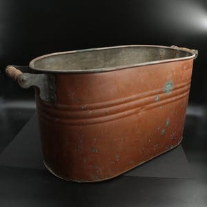 May include: An antique copper tub with wooden handles. The oval-shaped tub has a weathered, reddish-brown patina with visible signs of age and use. The handles are attached with metal brackets.