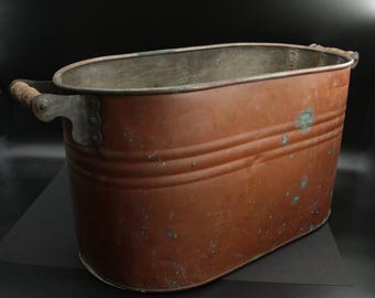 May include: An antique copper tub with wooden handles. The oval-shaped tub has a weathered, reddish-brown patina with visible signs of age and use. The handles are attached with metal brackets.