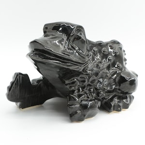 May include: A glossy black ceramic frog figurine with a textured, bumpy surface on its back and sides. The frog is depicted in a seated position with its mouth slightly open, showcasing a smooth, reflective finish across its entire body.
