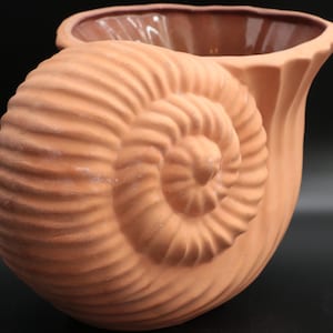 May include: A terracotta-colored ceramic vase with a seashell design. The vase features a spiral seashell shape on one side and a flared opening. The interior is a darker brown color.