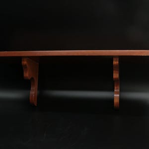 May include: A wooden wall shelf with a dark brown finish. The shelf has a rectangular top and two decorative brackets. The brackets have a curved, ornate design. The shelf is designed to be mounted on a wall for displaying items.