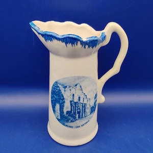 May include: White ceramic pitcher with a blue rim and handle. The pitcher features a blue and white image of the Alamo in San Antonio. The text "ALAMO - SAN ANTONIO" is below the image. The pitcher has a decorative, scalloped rim.