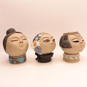 May include: Three ceramic face sculptures with closed eyes and pursed lips. The sculptures have tan skin tones and are adorned with various hairstyles and decorative elements, including flowers and headbands. Each sculpture sits atop a base with painted details.