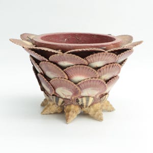 May include: A decorative bowl crafted from seashells, featuring a deep red interior and a layered design. The bowl's exterior is adorned with overlapping shells in shades of pink and cream, with a textured, beige base.