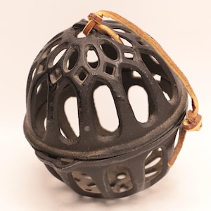 May include: A dark, spherical decorative object with an openwork design. The object is made of a dark metal and has a leather strap attached at the top. The sphere is divided into two halves. The openwork design features various shapes.