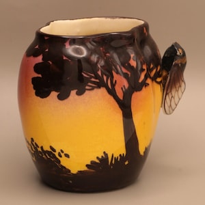 May include: A ceramic vase with a sunset design. The vase features a yellow and orange gradient with a black silhouette of a tree and foliage. A cicada-shaped handle is attached to the side. The vase has a unique, artistic design.