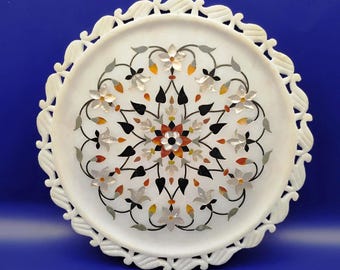 May include: A white marble decorative tray with a floral inlay design. The tray features a scalloped edge and a central floral pattern with black, orange, and white accents. The tray is round.