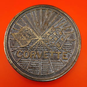 May include: A bronze-colored Corvette emblem with a circular design. The emblem features two crossed checkered flags above the word "CORVETTE". The background has a sunburst pattern. The emblem is set against an orange background.