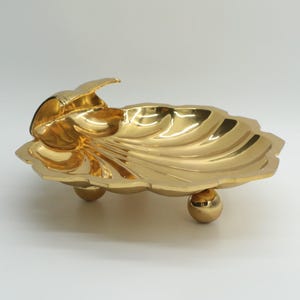 May include: A gold-colored decorative dish in the shape of a leaf, resting on three small ball feet. The dish has a textured, rippled surface and a curved, raised edge. The overall design is ornate and elegant.