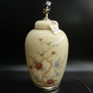 May include: Cream-colored ceramic table lamp with a floral design. The lamp features a silver base and a matching lid. The hand-painted design includes red, blue, and green flowers on a brown branch. The tag reads "Hand Decorated".