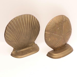 May include: Two brass bookends, one shaped like a seashell and the other like a sand dollar. The seashell bookend has a fan-like design with vertical ridges. The sand dollar bookend features a detailed pattern. Both have a rectangular base.
