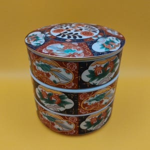 May include: A stacked, round ceramic container with a lid. The container is decorated with floral and landscape motifs in red, blue, and gold against a black and white background. The lid features a similar design. The container is likely for storage or display.