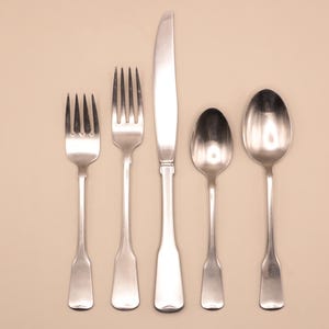 May include: A set of five silver-colored flatware pieces arranged vertically on a beige background. The set includes two forks, a knife, and two spoons. The utensils have a simple, classic design.