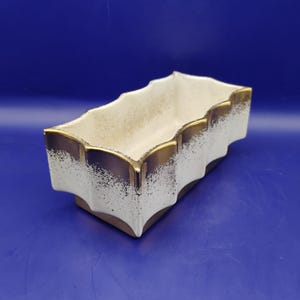 May include: Rectangular ceramic planter with a white and gold speckled design. The planter has a scalloped edge with gold trim. The interior is a light beige color. The planter sits on a brown base and is against a blue background.