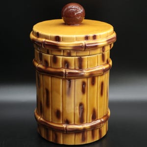 May include: A ceramic cookie jar with a barrel design. The jar is primarily tan with dark brown accents, including bands around the top, middle, and bottom. A small, round, dark brown knob sits atop the lid. The jar has a glossy finish.
