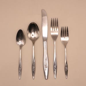 May include: A set of vintage silverware, including a knife, two forks, and two spoons, arranged on a neutral background. The silverware has a floral design on the handles and a shiny, reflective surface.