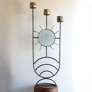 May include: A tall, black metal candelabra with three candle holders. The design includes a frosted glass sun in the center and curved lines at the base. The candle holders are gold-colored.