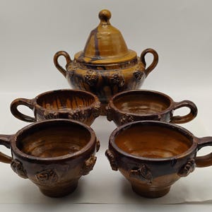 May include: A ceramic tea set with a sugar bowl and four cups. The set is glazed in shades of brown and tan, with a textured surface and grape cluster detailing. The sugar bowl has a lid and two handles.