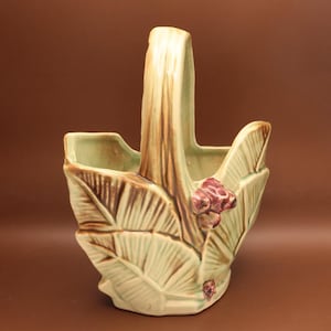 May include: A ceramic basket with a handle, featuring a leaf design in shades of green and brown. The basket has a textured surface and is adorned with small, reddish-purple floral accents. The handle is a light brown color.