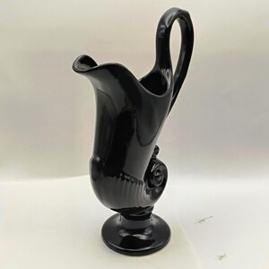 May include: A glossy black vase with a unique, curved handle and a flared spout. The vase has a seashell-like design on its body and sits on a round base. The overall shape is elegant and decorative.