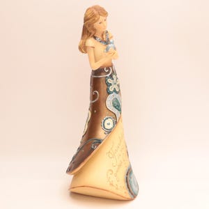 May include: A decorative figurine of a woman holding a small bird. The figurine has long brown hair and is wearing a tan top and a long brown skirt with blue and white floral designs. The base of the figurine is tan with script that reads "Friendship is a gift".