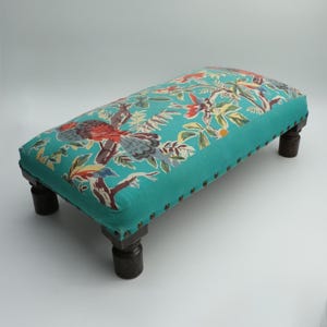 May include: A rectangular footstool with a teal fabric top featuring a colorful bird and floral pattern. The edges are trimmed with small metal studs. The dark wooden legs are approximately 6 inches tall.