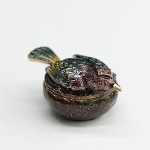 May include: A decorative bird-shaped trinket box. The bird has iridescent blue, green, and purple feathers, a gold beak, and a small crown. The base of the box is a speckled dark brown color with gold flecks.