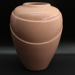 May include: A peach-colored ceramic vase with a rounded body and a narrow neck. The vase features two curved, horizontal ridges that wrap around the body. The vase has a glossy finish and is set against a dark background.