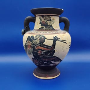 May include: A cream-colored ceramic vase with black handles and a dark brown base. The vase features ancient Greek-style illustrations in black and red, depicting figures playing instruments and wearing traditional attire. The vase is approximately 8 inches tall.