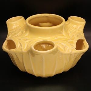May include: A yellow ceramic flower frog with multiple openings for arranging flowers. The frog has a central circular opening surrounded by smaller openings and cylindrical tubes. The exterior features a ribbed design and leaf-like details.