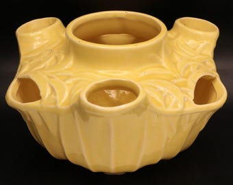 May include: A yellow ceramic flower frog with multiple openings for arranging flowers. The frog has a central circular opening surrounded by smaller openings and cylindrical tubes. The exterior features a ribbed design and leaf-like details.