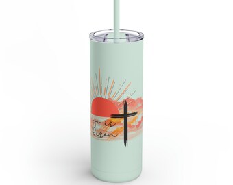 May include: A light green stainless steel tumbler with a sunrise and cross design. The tumbler has the text "He is risen" on it.