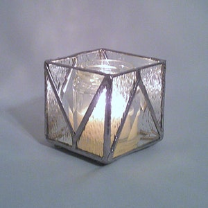 Pode incluir: Um porta-velas de vidro transparente com um design geométrico. O porta-velas é feito de vidro colorido e tem uma forma quadrada. Uma vela está a arder dentro do porta-velas.