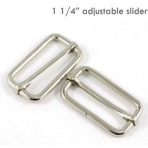 May include: Two silver-colored, rectangular adjustable sliders. The text "1 1/4" adjustable slider" is above the sliders. These metal sliders are used for adjusting straps on bags, clothing, or other accessories.