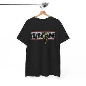May include: Black t-shirt with the word "TRIC" in white outlined letters, a yellow lightning bolt, and red and yellow horizontal lines. The shirt is hanging on a wooden hanger.