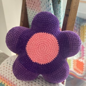 Puede incluir: Una almohada de color morado en forma de flor hecha a crochet con un centro rosa. La almohada est&aacute; sobre una manta de crochet blanca y de colores pastel.