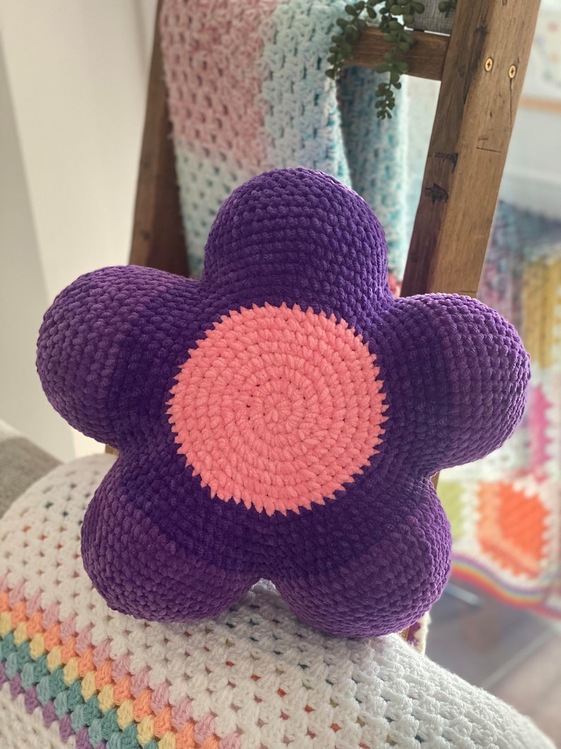 Puede incluir: Una almohada de color morado en forma de flor hecha a crochet con un centro rosa. La almohada est&aacute; sobre una manta de crochet blanca y de colores pastel.