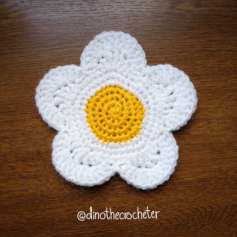 Może przedstawiać: Biały kwiat na szydełku z ż&oacute;łtym środkiem. Kwiat leży na drewnianej powierzchni. @dinothecrocheter