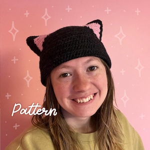 May include: A black crocheted cat ear hat with pink ears. The hat is worn by a person with brown hair and a light pink background.
